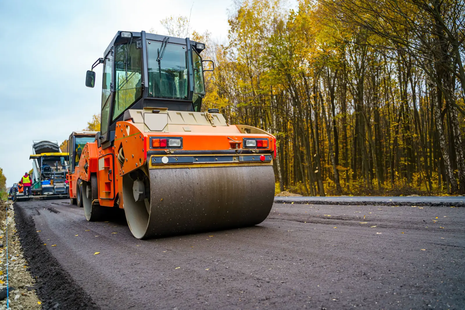 Road roller compacting fresh asphalt on a two-lane road, paving crew working in the background
