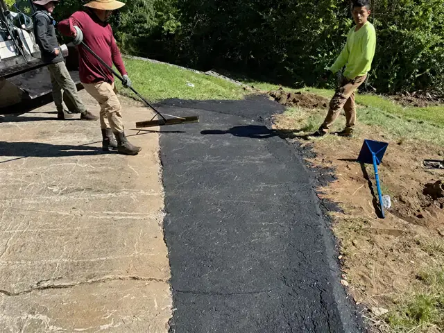 Two workers raking hot asphalt at the edge of a concrete apron